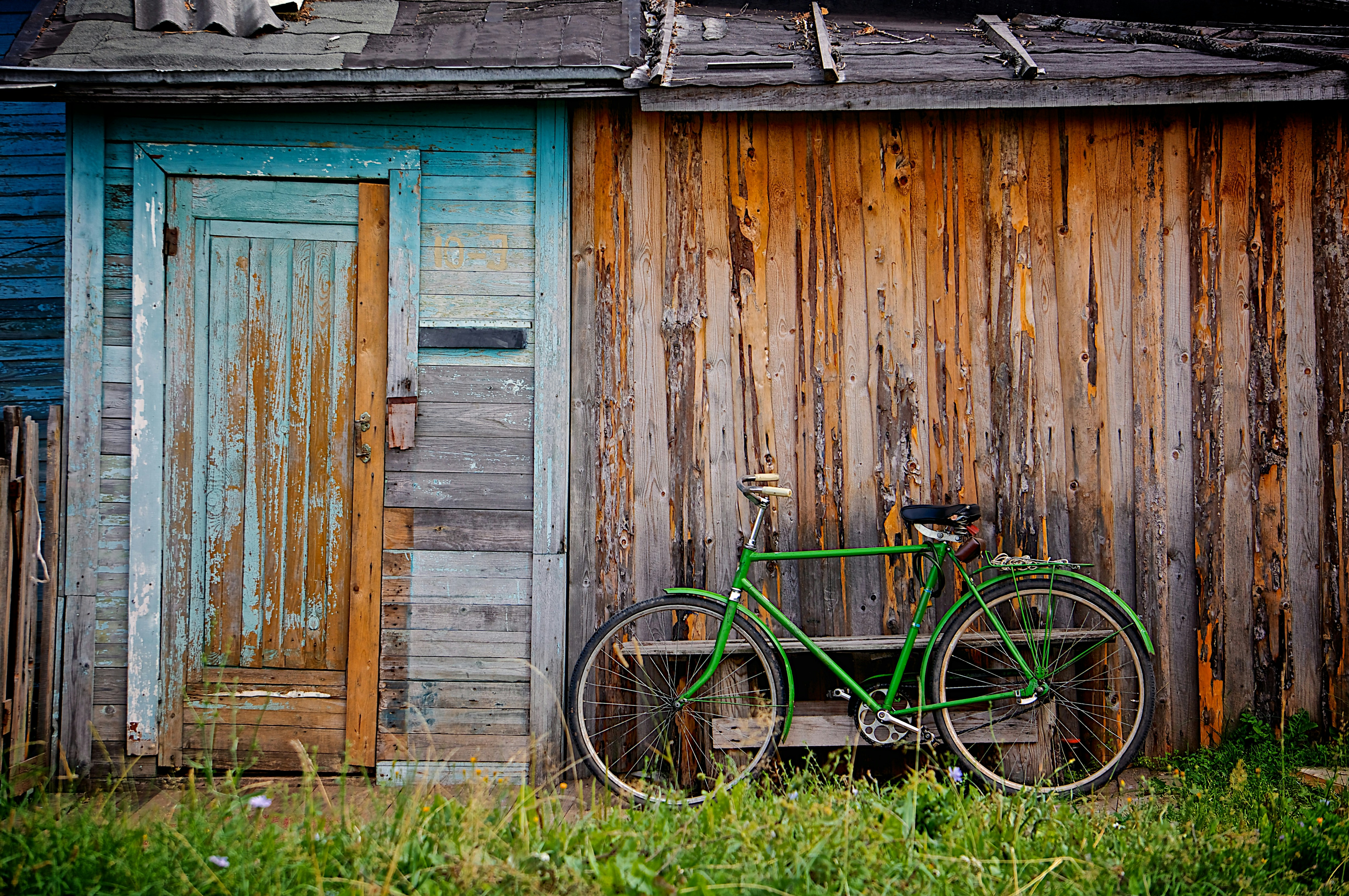 An old-timey green bicycycle leaning against an old derelict wooden shed. The paint is flaking off but it was once a blue shed.